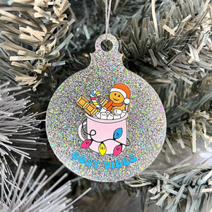 Close up of a white Christmas tree displaying a silver glitter bauble featuring retro text reading 'cosy vibes'. The image shows a mug of hot chocolate with marshmallows and a gingerbread man wearing a Santa hat. A candy cane in the mug and lights around the mug are the colours of the pansexual pride flag.