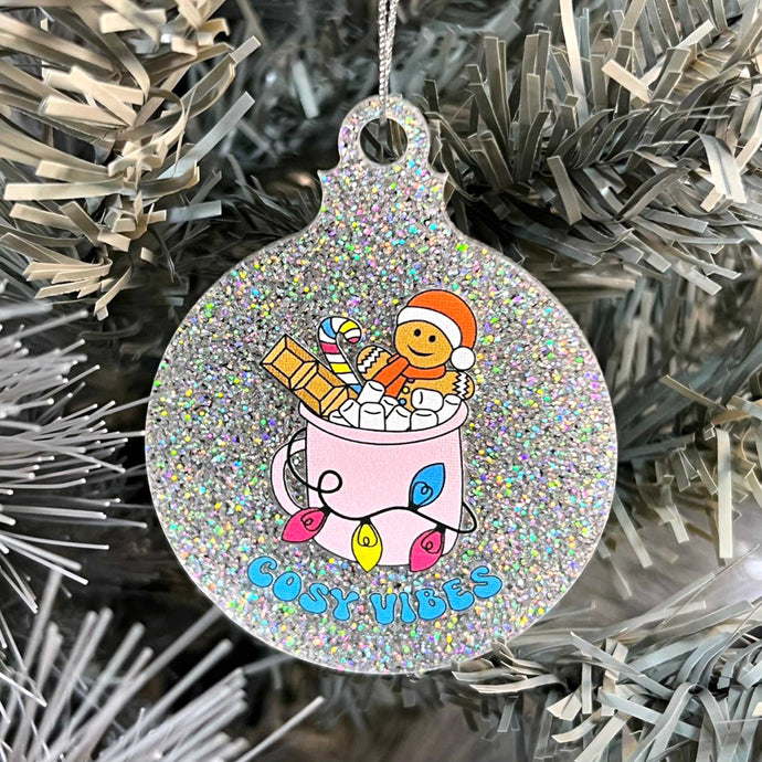 Close up of a white Christmas tree displaying a silver glitter bauble featuring retro text reading 'cosy vibes'. The image shows a mug of hot chocolate with marshmallows and a gingerbread man wearing a Santa hat. A candy cane in the mug and lights around the mug are the colours of the pansexual pride flag.