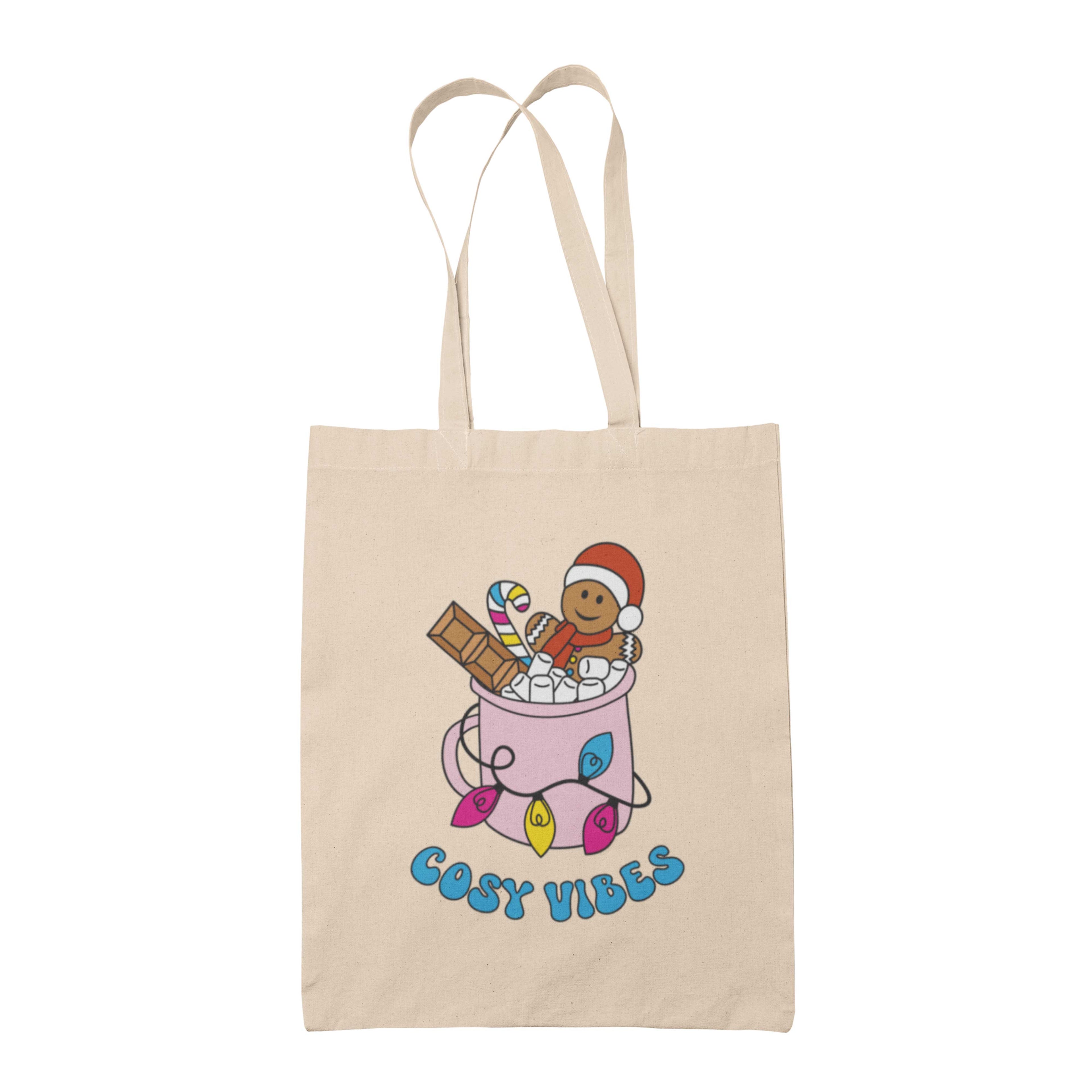 Natural tote bag featuring retro text reading 'cosy vibes'. The image shows a mug of hot chocolate with marshmallows and a gingerbread man wearing a Santa hat. A candy cane in the mug and lights around the mug are the colours of the pansexual pride flag.