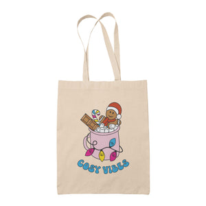 Natural tote bag featuring retro text reading 'cosy vibes'. The image shows a mug of hot chocolate with marshmallows and a gingerbread man wearing a Santa hat. A candy cane in the mug and lights around the mug are the colours of the pansexual pride flag.