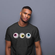 Load image into Gallery viewer, A man wearing a dark heather grey t-shirt stands against a plain grey background. The shirt features a design of three cartoon eyeballs with detailed red veins, each eye representing different pride colours. The left eye has a pink iris, the middle eye has a yellow iris, and the right eye has a blue iris, corresponding to the Pansexual pride flag colours. The man has a short haircut and is looking directly at the camera with a neutral expression.