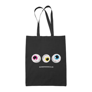 A black tote bag with three cartoonish eyeballs in a row, each with different coloured irises: pink, yellow, and blue. The design is finished with the text "RAINBOWANDCO.UK" beneath the eyeballs.