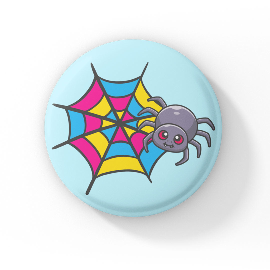 A circular badge showcasing a cute grey spider with pink eyes on a web coloured in pink, yellow, and blue, representing the pansexual pride flag. The badge has a light blue background.