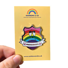 Load image into Gallery viewer, White hand holding up an enamel pin on a yellow backing card. The pin is in the shape of a shield with a banner across it&#39;s front. Banner reads Pridesmate. The background of the pin is the Gilbert Baker pride flag and there is a pair of cheersing champagne glasses in the centre. The champage and banner are filled with white glitter.