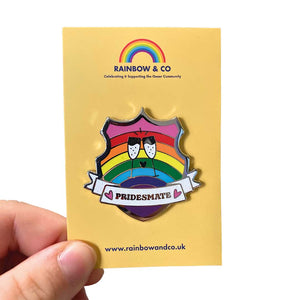 White hand holding up an enamel pin on a yellow backing card. The pin is in the shape of a shield with a banner across it's front. Banner reads Pridesmate. The background of the pin is the Gilbert Baker pride flag and there is a pair of cheersing champagne glasses in the centre. The champage and banner are filled with white glitter.