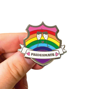 White hand holding up an enamel pin in the shape of a shield with a banner across it's front. Banner reads Pridesmate. The background of the pin is the Gilbert Baker pride flag and there is a pair of cheersing champagne glasses in the centre. The champage and banner are filled with white glitter.