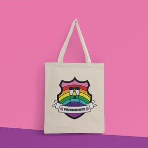 Natural coloured tote bag hanging from a peg on a pink wall. The bag features a shield with a banner across it's front. Banner reads Pridesmate. The background of the shield is the Gilbert Baker pride flag and there is a pair of clinking champagne flutes in the centre.