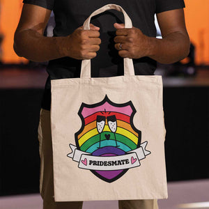 Close up of a black person holding a natural coloured tote bag featuring a shield with a banner across it's front. Banner reads Pridesmate. The background of the shield is the Gilbert Baker pride flag and there is a pair of clinking champagne flutes in the centre.