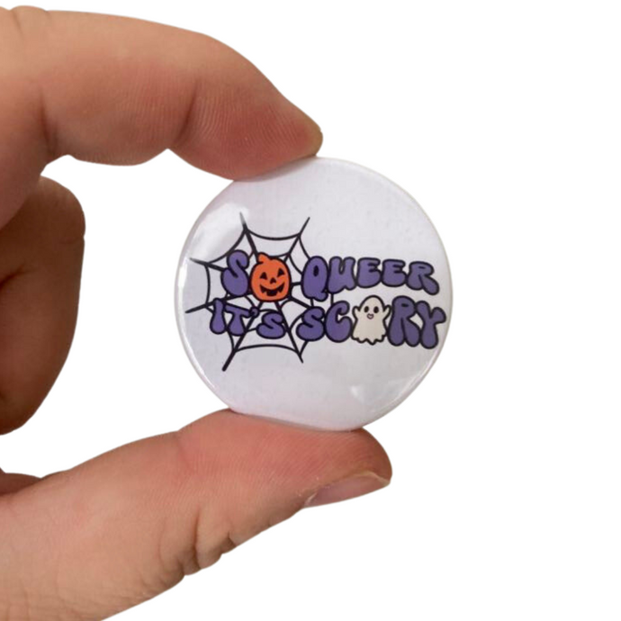 A white 38mm badge with text reading 'So Queer It's Scary' with images of a pumpkin, ghost, and spiders web