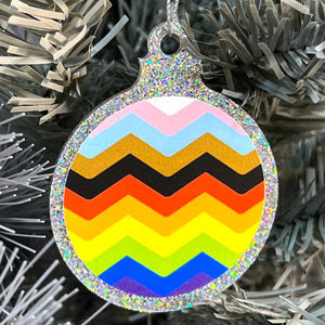Close up of a white Christmas tree displaying a silver glitter bauble featuring a classic zig zag design in the colours of the progress pride flag.