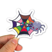 Load image into Gallery viewer, A hand holding a cute spider sticker with a progress pride web design. The web displays a variety of colours, including brown, black, pink, blue, red, orange, yellow, and green, representing the progress pride flag.