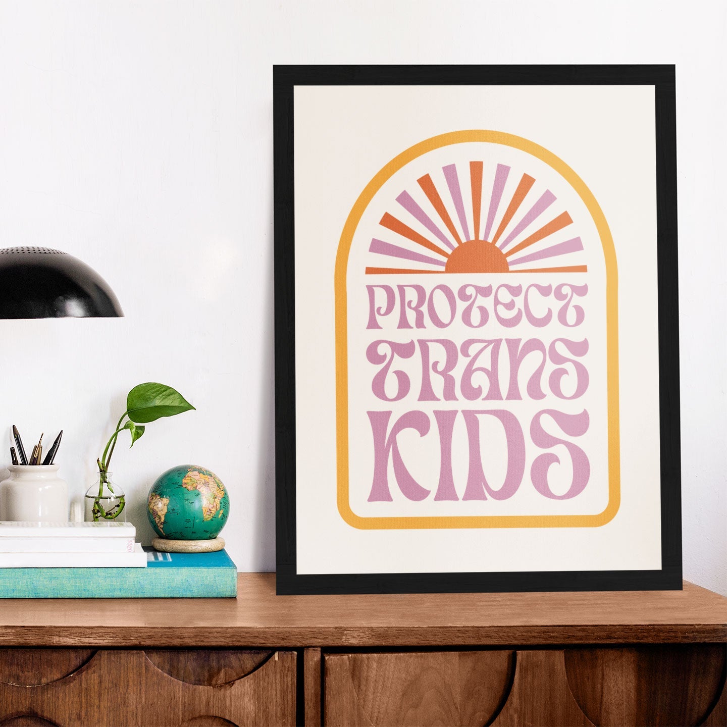 A framed Protect Trans Kids print featuring a retro arch design with a pink and orange sunburst, displayed on a wooden sideboard with books, a globe, and a black desk lamp.