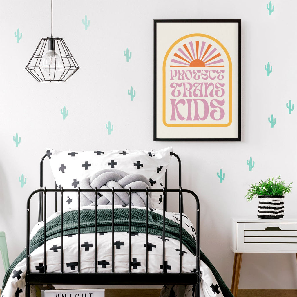 A framed Protect Trans Kids print hanging on the wall above a black metal bed with green and white bedding, surrounded by minimalist cactus wall decals.