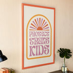 A Protect Trans Kids print in a modern workspace, framed with an orange border, resting on a wooden desk next to a black lamp, books, and a laptop.