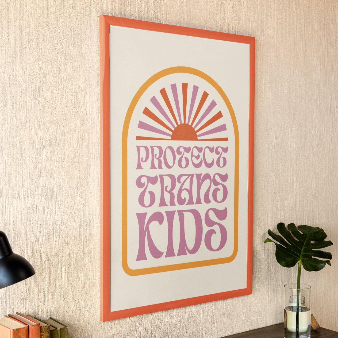 A Protect Trans Kids print in a modern workspace, framed with an orange border, resting on a wooden desk next to a black lamp, books, and a laptop.