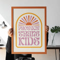 A person sits in a modern space holding a framed Protect Trans Kids print. The design features warm pastel hues with a rising sun illustration and retro typography.