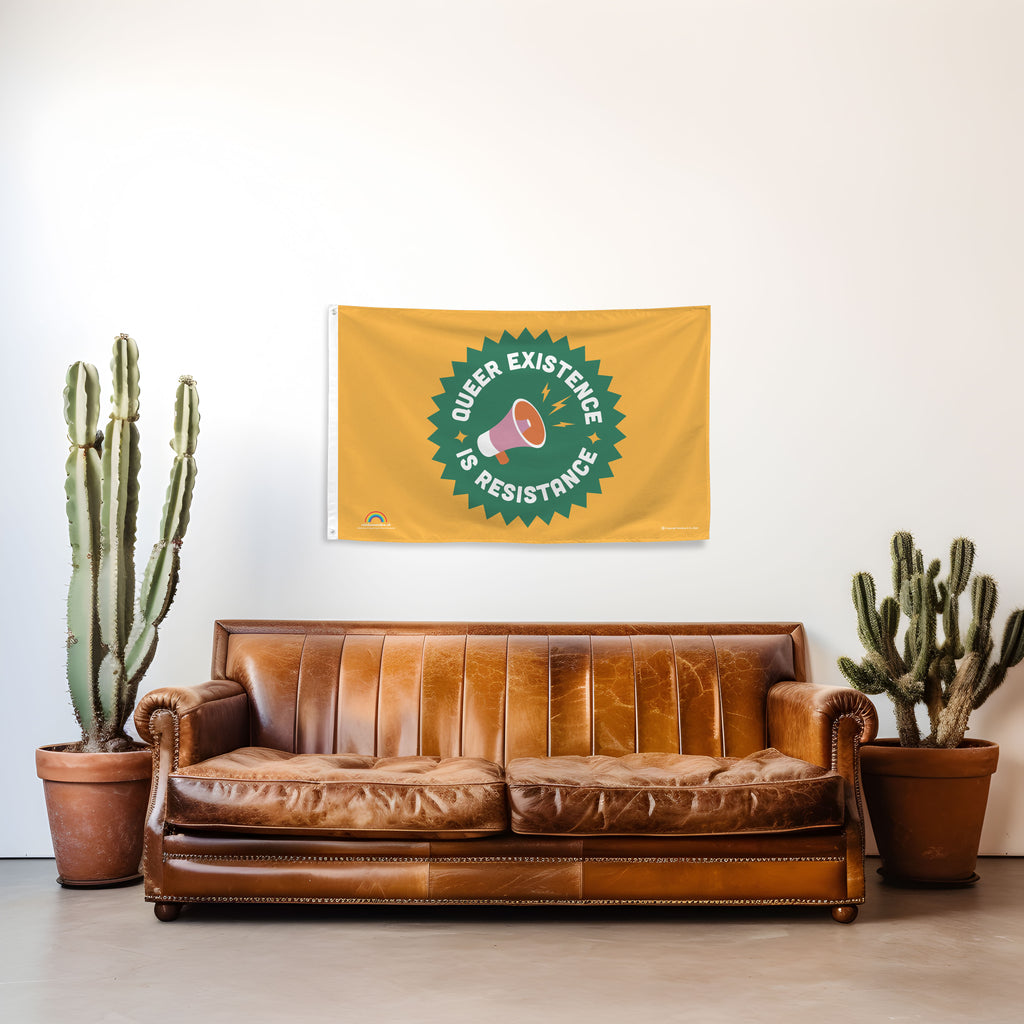 Flag with 'Queer Existence is Resistance' design on a wall above a brown leather sofa with cacti on either side.