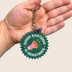 A hand holds a round, jagged-edge acrylic keyring in front of a light beige background. The keyring reads “QUEER EXISTENCE IS RESISTANCE” in bold white text, circling a pink and orange megaphone with yellow lightning bolts around it. The keyring hangs from a silver chain and includes a small clear charm with the Rainbow & Co logo in dark blue.
