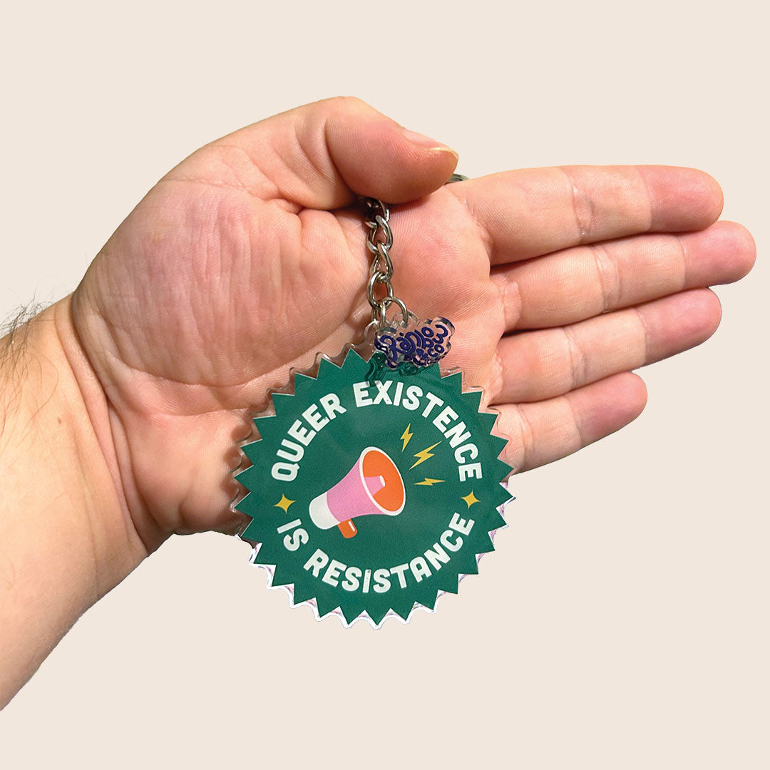 A “Queer Existence is Resistance” keyring is held by a hand, with the keyring positioned more centrally and higher up. The light beige background and Rainbow & Co charm are visible again, with the same bold white text and megaphone design.