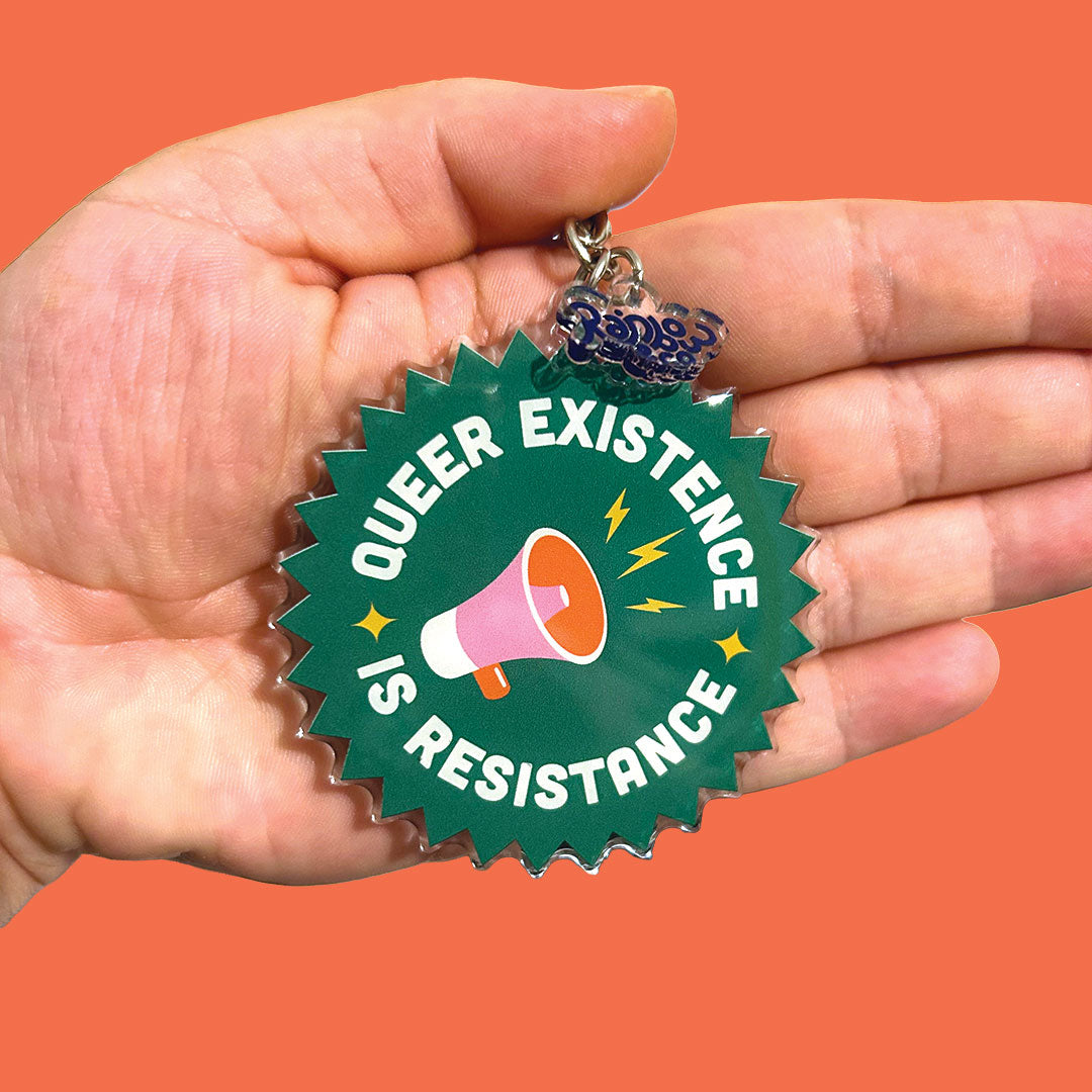 The “Queer Existence is Resistance” keyring is shown against a bright orange background, held in a palm-facing-forward position. The vivid green of the keyring contrasts with the background, and the white lettering, megaphone, and lightning bolts are clearly visible.