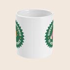 A white ceramic mug shown with the handle facing away, displaying part of a bold green starburst graphic with a visible pink and orange megaphone. The image shows how the graphic is repeated on each side of the mug. The background is a soft beige.