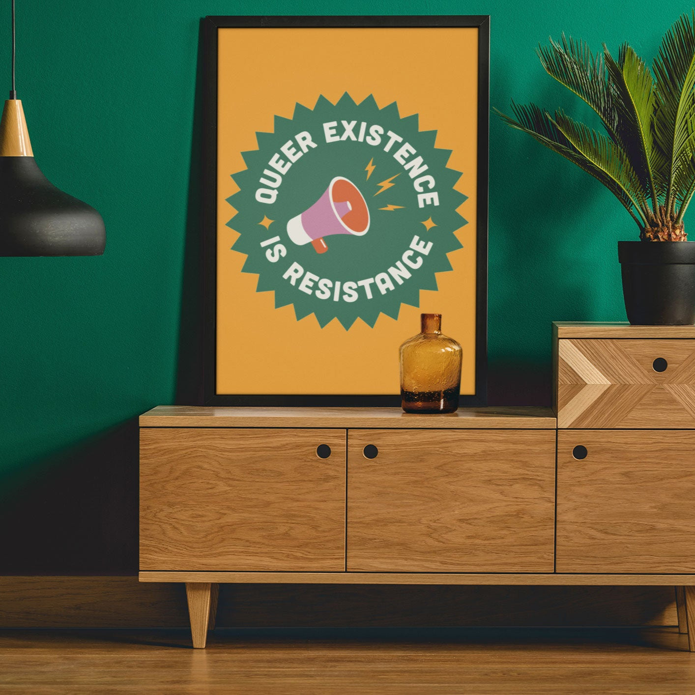 A framed Queer Existence is Resistance print with a bold yellow and green design, featuring a megaphone illustration, displayed on a modern wooden sideboard with a black pendant lamp and a potted plant.