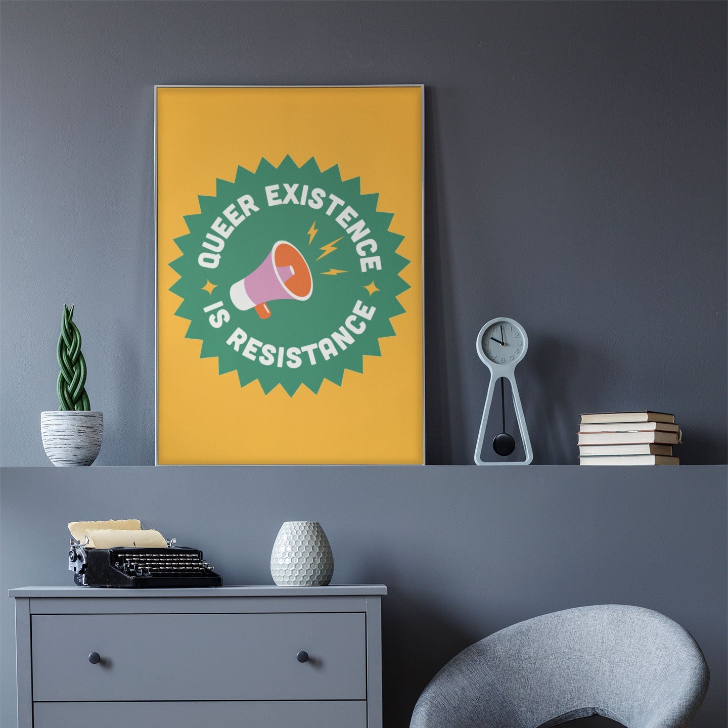A Queer Existence is Resistance print hanging on a grey wall above a minimalist chest of drawers with a vintage typewriter and books