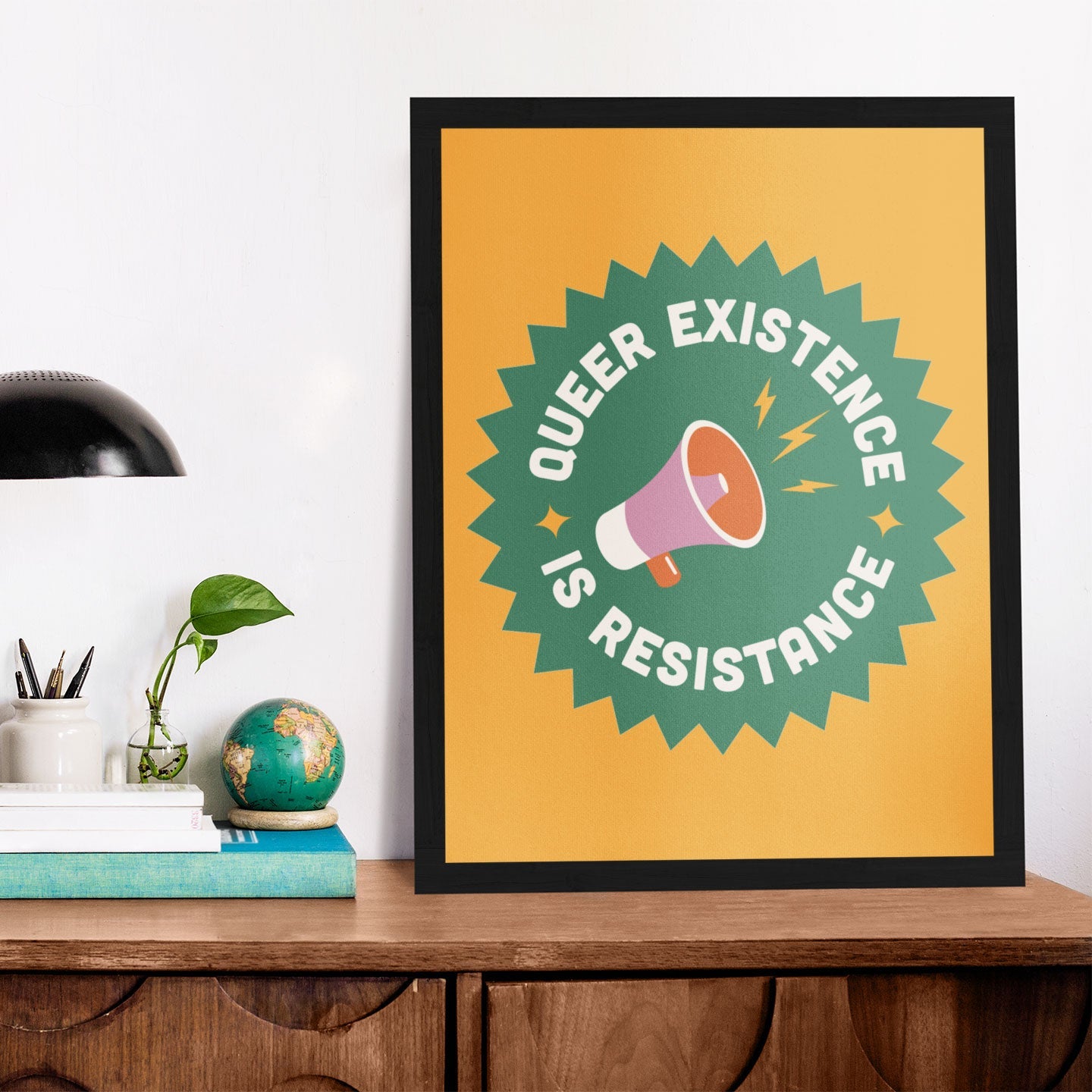 A framed Queer Existence is Resistance print leaning against a white wall on a wooden desk, accompanied by a black lamp, books, and a small potted plant.