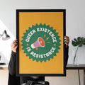 A framed Queer Existence is Resistance print is held by a seated person, featuring a bold yellow and green colour scheme with a megaphone icon and striking typography.