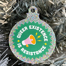 Load image into Gallery viewer, Close up of a white Christmas tree displaying a silver glitter bauble featuringthe text Queer Existence is Resistance on a green background with a graphic of a megaphone in pink and orange.