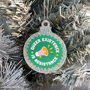 Close up of a white Christmas tree displaying a silver glitter bauble featuringthe text Queer Existence is Resistance on a green background with a graphic of a megaphone in pink and orange.