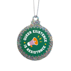A silver glitter bauble against a white background featuring the text Queer Existence is Resistance on a green background with a graphic of a megaphone in pink and orange. It is held up by a silver string.