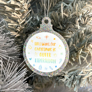 Close up of a white Christmas tree displaying a silver glitter bauble featuring the text All I Want for Christmas is QUEER LIBERATION surrounded by Christmas icons such as candy canes and stockings, all in rainbow pride colours.