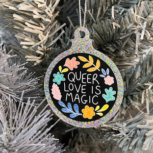 Close up of a white Christmas tree displaying a silver glitter bauble featuring the text Queer Love is Magic and hand drawn illustrations of flowers in rainbow colours.
