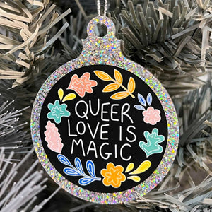 Close up of a white Christmas tree displaying a silver glitter bauble featuring the text  Queer Love is Magic and hand drawn illustrations of flowers in rainbow colours.