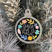 Load image into Gallery viewer, Close up of a white Christmas tree displaying a silver glitter bauble featuring the text Queer Love is Magic and hand drawn illustrations of flowers in rainbow colours.