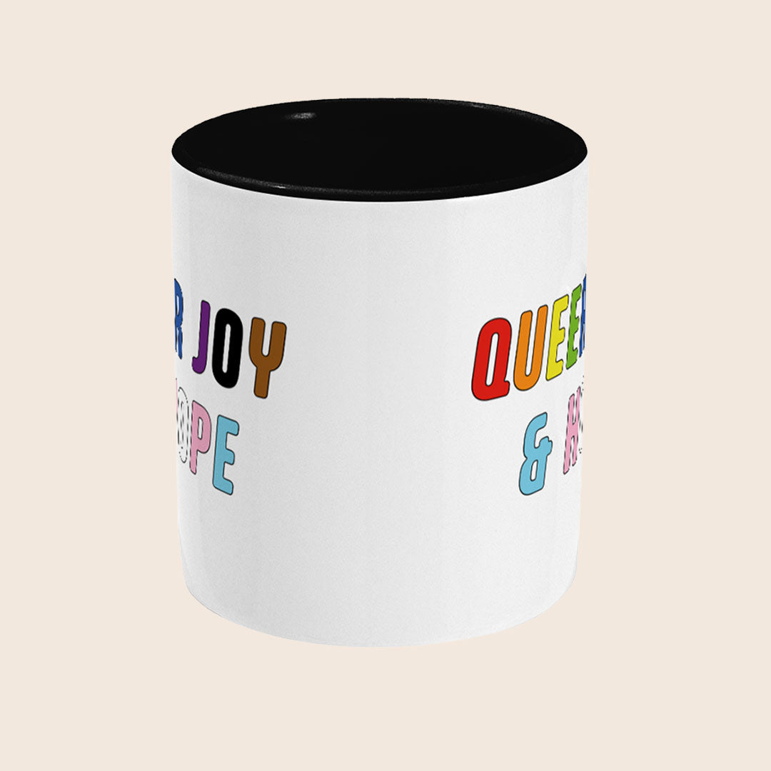 White mug with black interior featuring the colourful words “Queer Joy & Hope” wrapping around both sides.