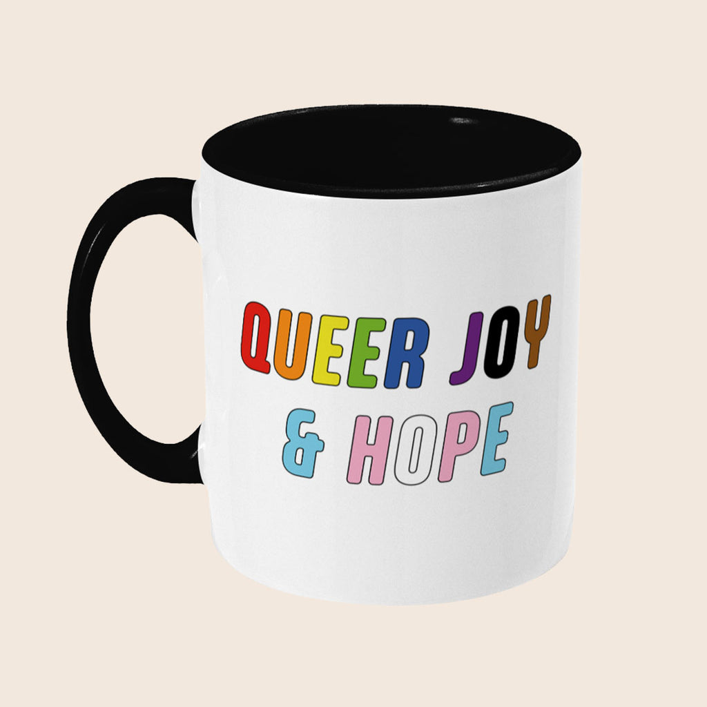 White mug with black handle and interior featuring the colourful words “Queer Joy & Hope” on the front.
