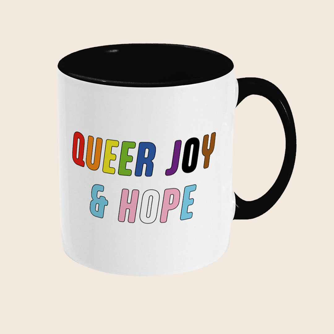 White mug with black handle and interior featuring the colourful words “Queer Joy & Hope” on the front.