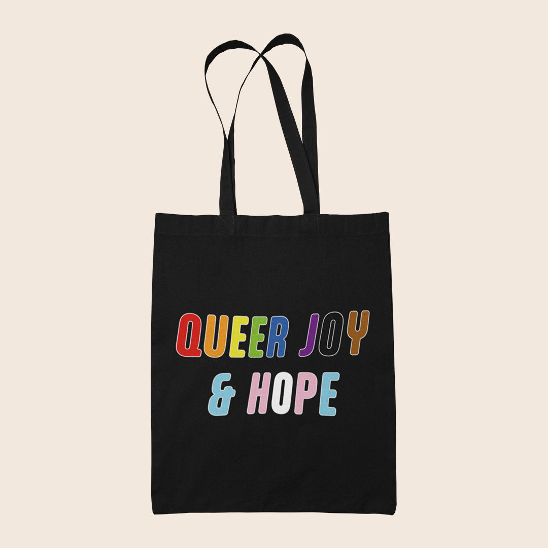 Black tote bag featuring colourful text reading “Queer Joy & Hope” in rainbow and trans Pride-inspired colours.