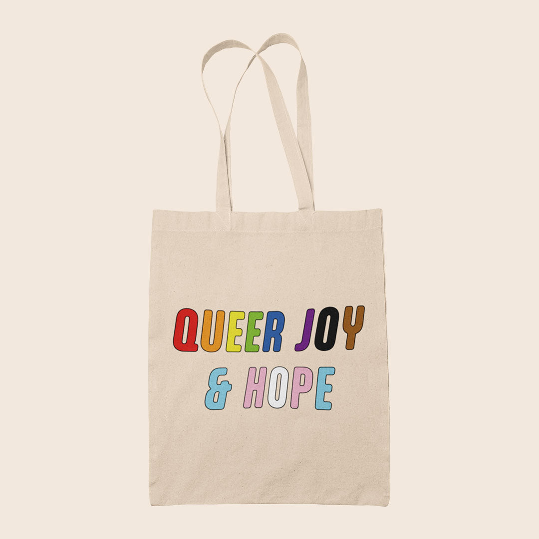 Natural cotton tote bag featuring colourful text reading “Queer Joy & Hope” in rainbow and trans Pride-inspired colours.