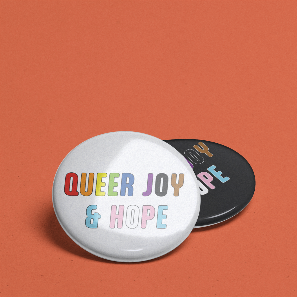 Two round Queer Joy & Hope badges overlapping on an orange background. A white badge with colourful lettering sits in front of a black badge featuring the same design.