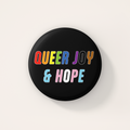Black round 38mm badge featuring the words "Queer Joy & Hope" in bold, multicoloured lettering inspired by the rainbow and transgender Pride flags.