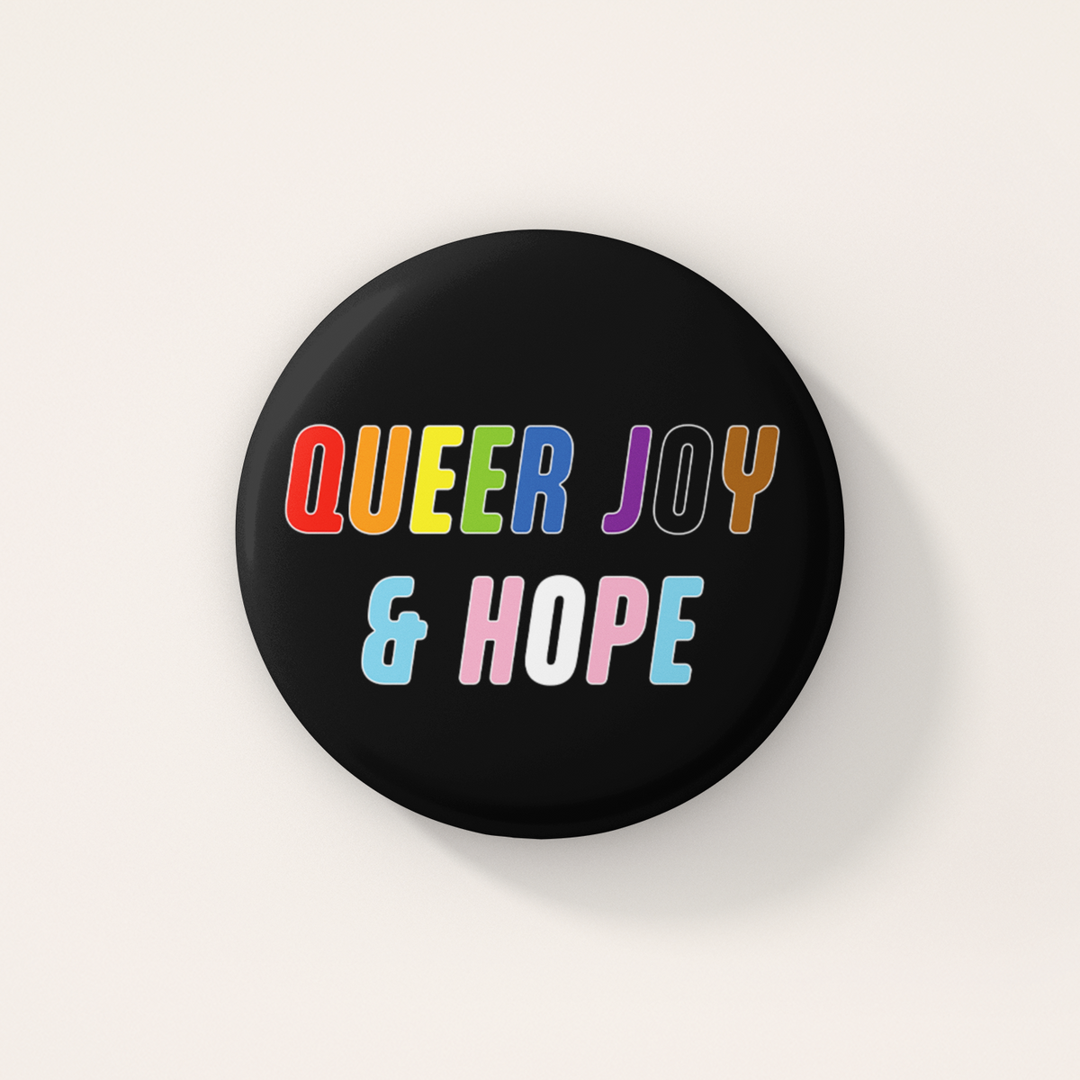 Black round 38mm badge featuring the words "Queer Joy & Hope" in bold, multicoloured lettering inspired by the rainbow and transgender Pride flags.
