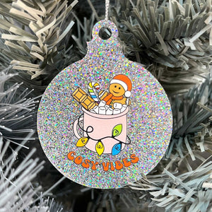 Close up of a white Christmas tree displaying a silver glitter bauble featuring retro text reading 'cosy vibes'. The image shows a mug of hot chocolate with marshmallows and a gingerbread man wearing a Santa hat. A candy cane in the mug and lights around the mug are the colours of the rainbow pride flag.