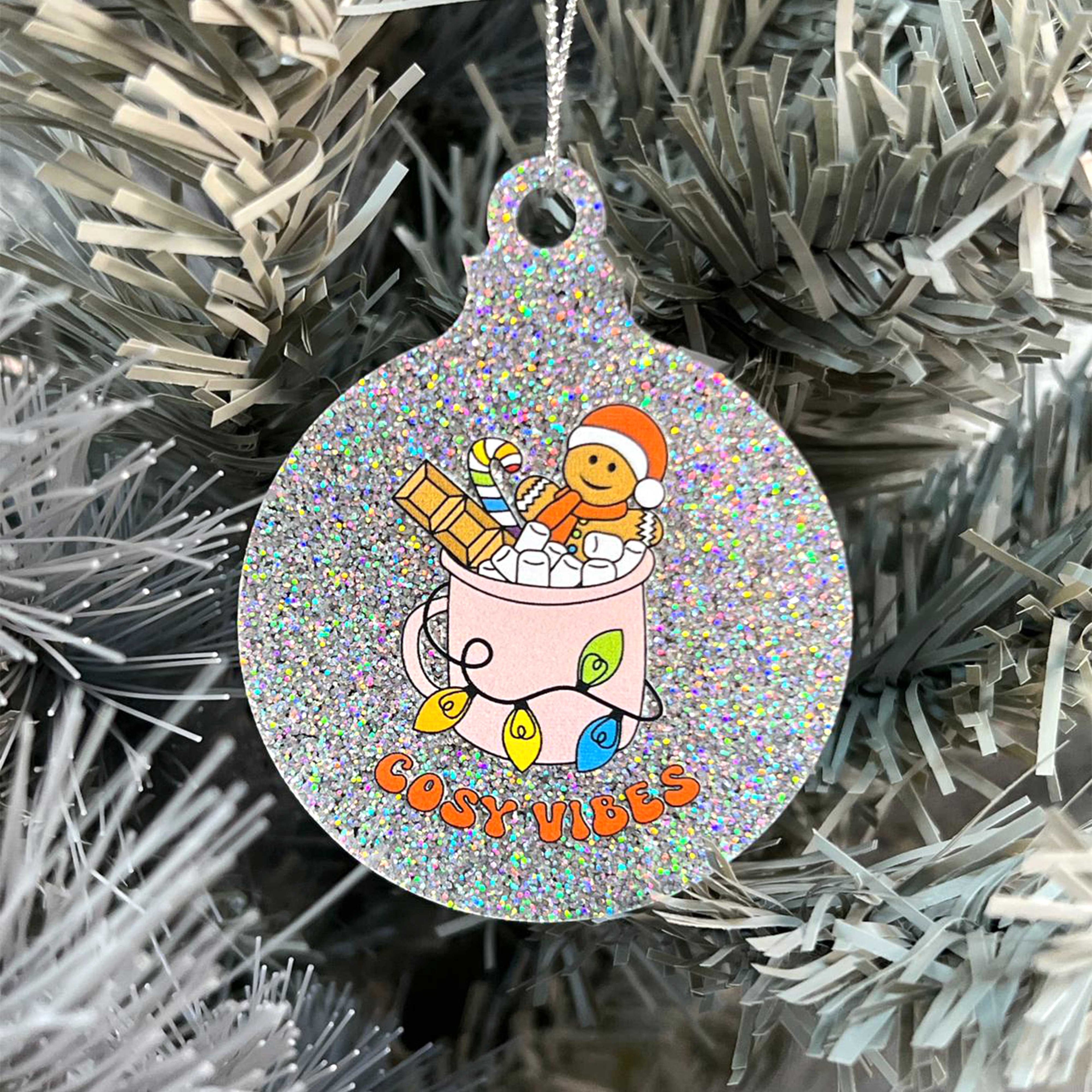 Close up of a white Christmas tree displaying a silver glitter bauble featuring retro text reading 'cosy vibes'. The image shows a mug of hot chocolate with marshmallows and a gingerbread man wearing a Santa hat. A candy cane in the mug and lights around the mug are the colours of the rainbow pride flag.