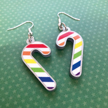 Load image into Gallery viewer, Silver glitter candy cane dangle earrings lay on a green star paper background. The stripes on the candy cane are those of the Rainbow flag; red, orange, yellow, green, blue, purple.