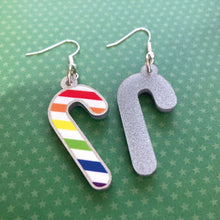 Load image into Gallery viewer, Silver glitter candy cane dangle earrings lay on a green star paper background. The stripes on the candy cane are those of the Rainbow flag; red, orange, yellow, green, blue, purple. One of the earrings is turned over to show the reverse, which is plain silver glitter.