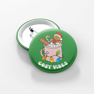 Close up of a green 38mm badge featuring retro text reading 'cosy vibes'. The image shows a mug of hot chocolate with marshmallows and a gingerbread man wearing a Santa hat. A candy cane in the mug and lights around the mug are the colours of the rainbow pride flag.