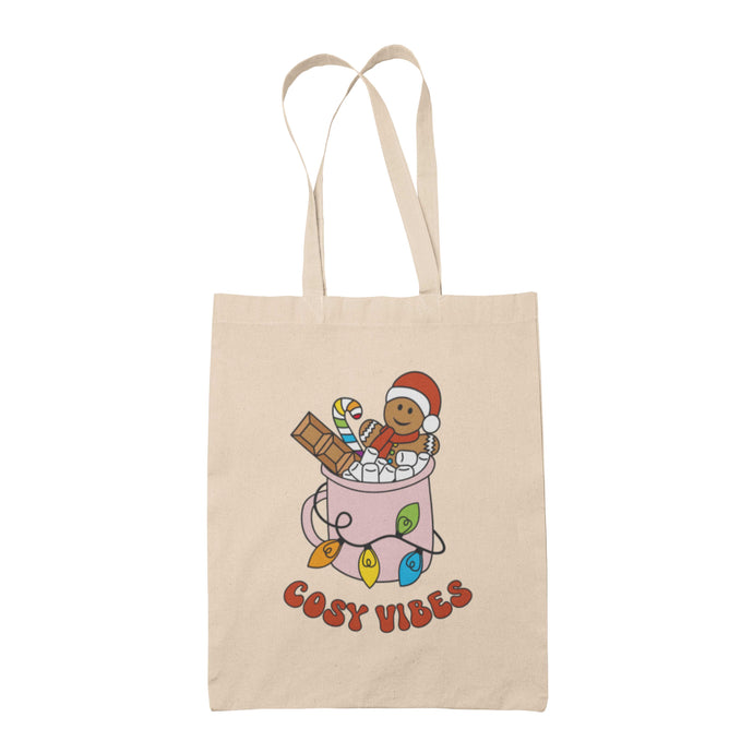 Natural tote bag featuring retro text reading 'cosy vibes'. The image shows a mug of hot chocolate with marshmallows and a gingerbread man wearing a Santa hat. A candy cane in the mug and lights around the mug are the colours of the rainbow pride flag.